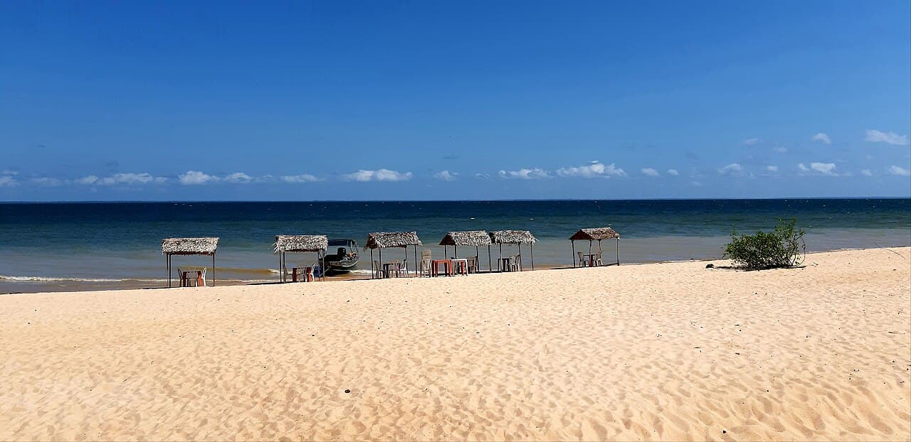 Faixa larga de areia clara de praia de rio em Alter do Chão, com meia dúzia de quiosques de palha e cadeiras plásticas alinhados na beira da água esverdeada, sob céu azul limpo