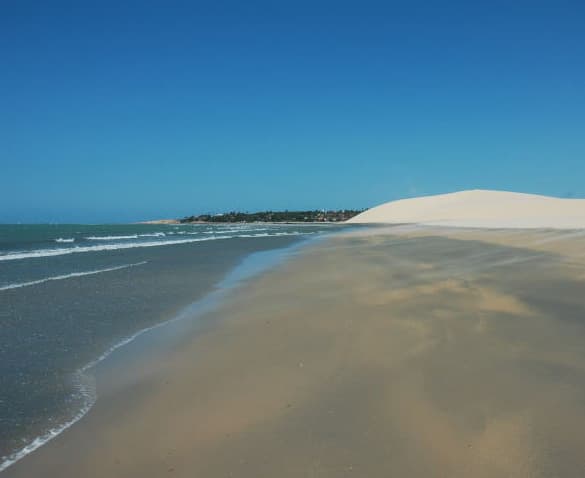 Praia larga de areia úmida em Jericoacoara, com o mar quebrando à esquerda, uma duna branca alta à direita e a vila ao fundo sobre a faixa de vegetação baixa