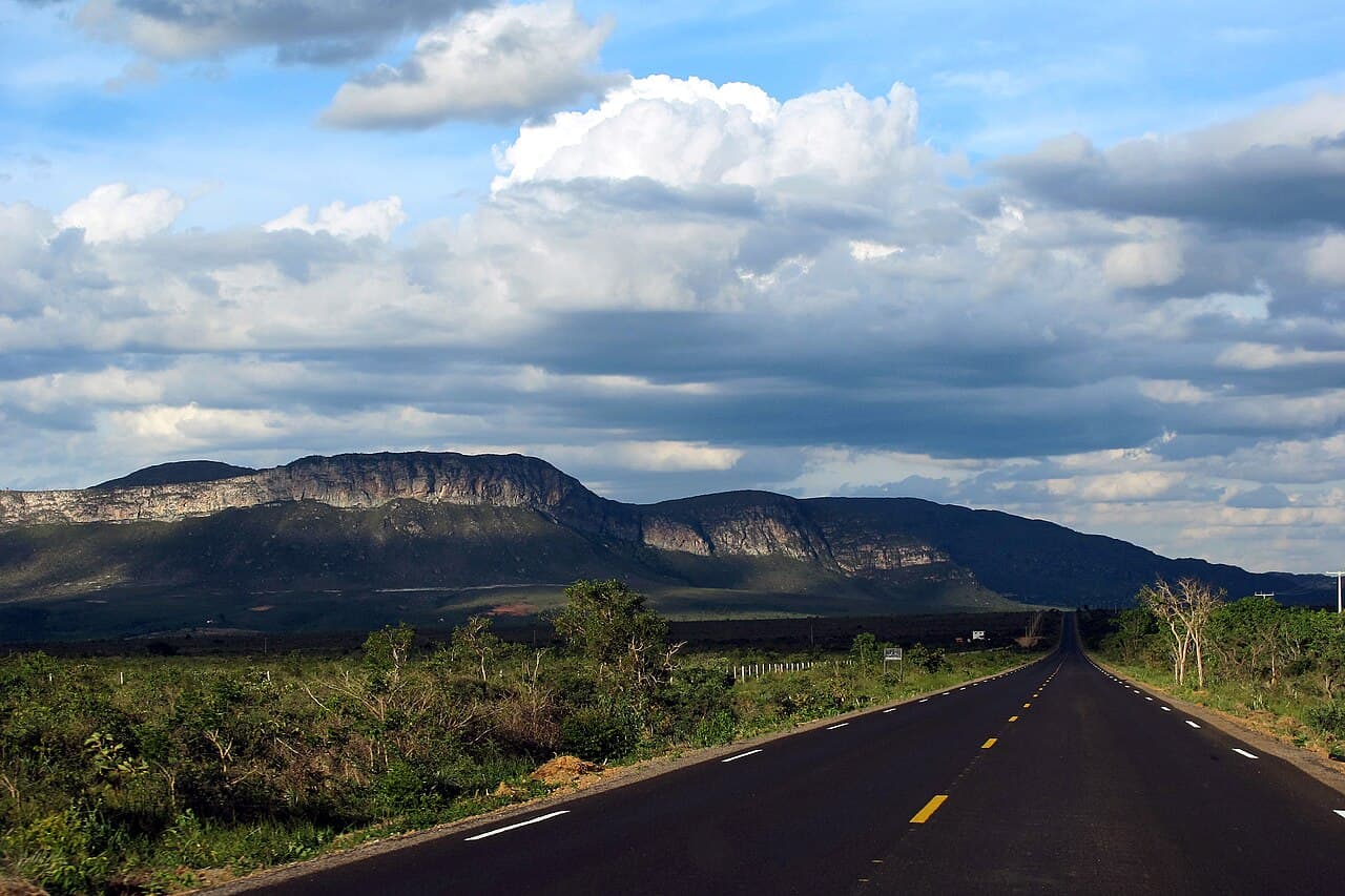 Rodovia asfaltada reta cortando a caatinga verde da Chapada Diamantina, com o paredão de uma serra de topo plano no horizonte e nuvens altas acima