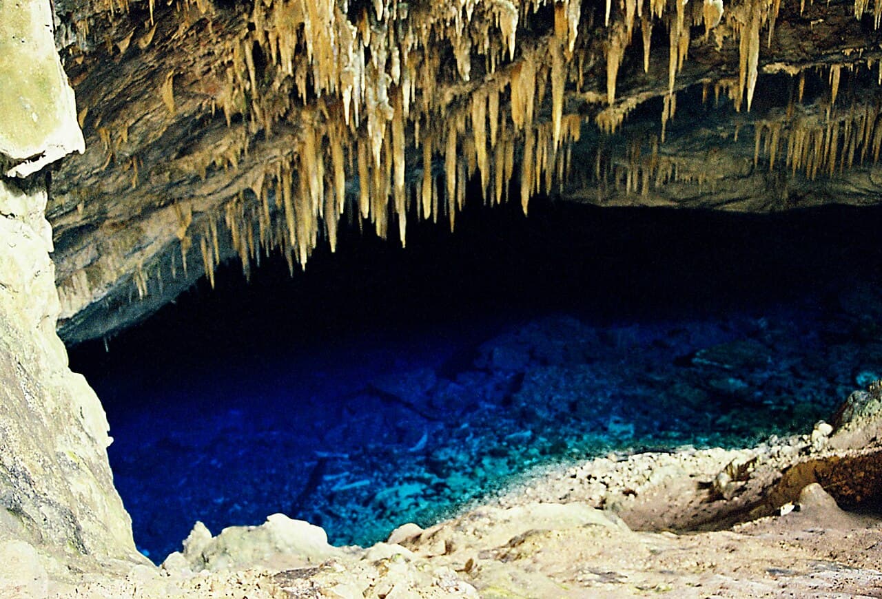 Interior da Gruta do Lago Azul, com estalactites alaranjadas no teto e o lago azul-escuro iluminado no fundo da caverna