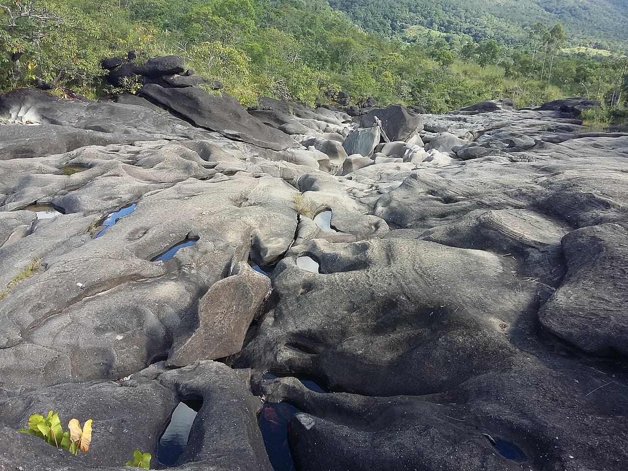 Leito de rocha cinza do Vale da Lua esculpido em bacias e canais, com pouca água nos poços e cerrado verde ao fundo