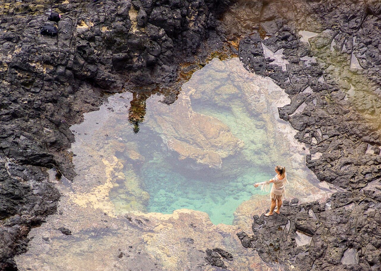 Piscina natural de água esverdeada entre rochas escuras em Fernando de Noronha, vista de cima, com uma pessoa em pé na borda dando noção de escala