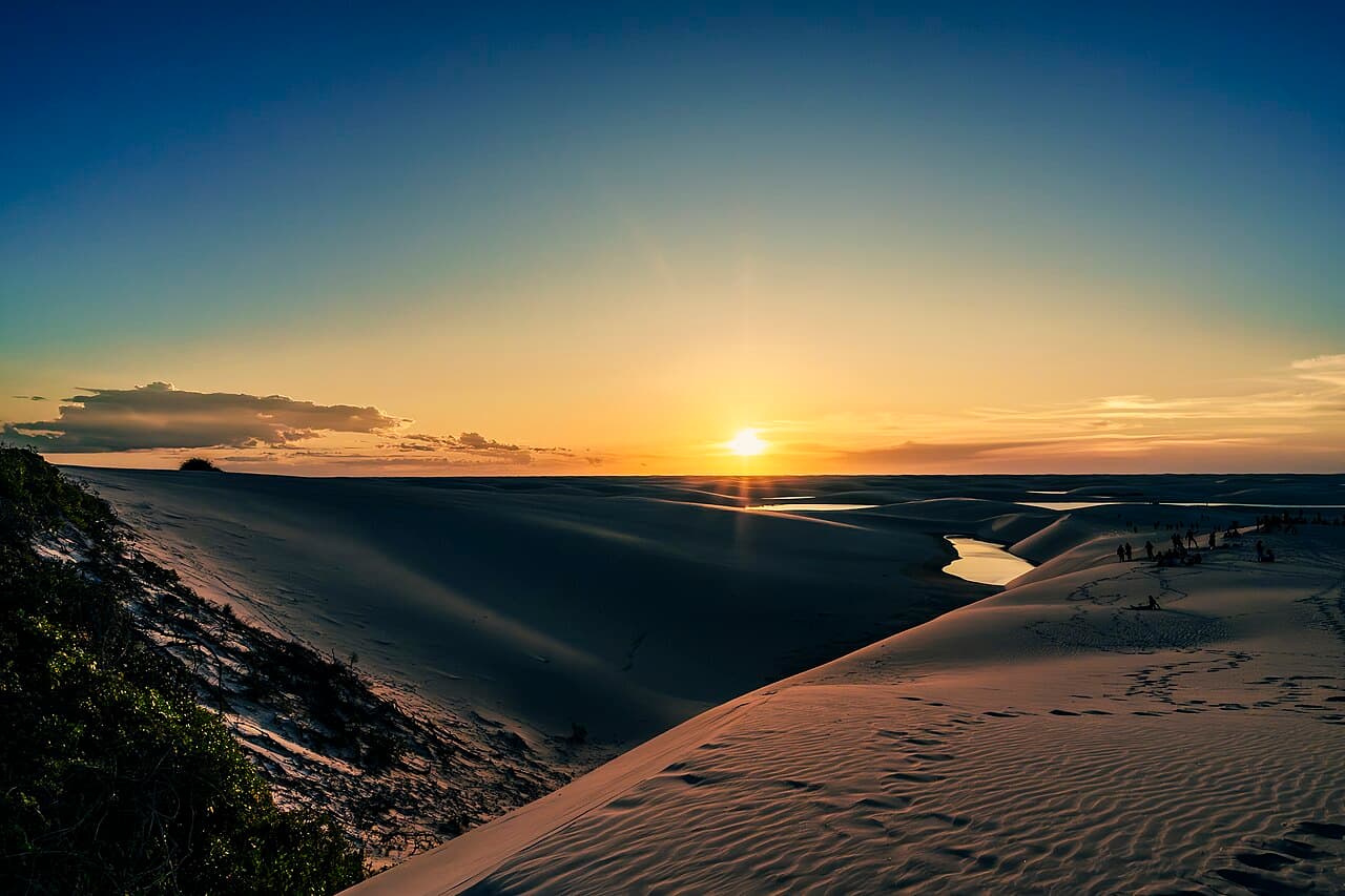 Pôr do sol sobre as dunas dos Lençóis Maranhenses, com lagoas refletindo a luz ao fundo e um grupo de pessoas de pé na crista da duna à direita