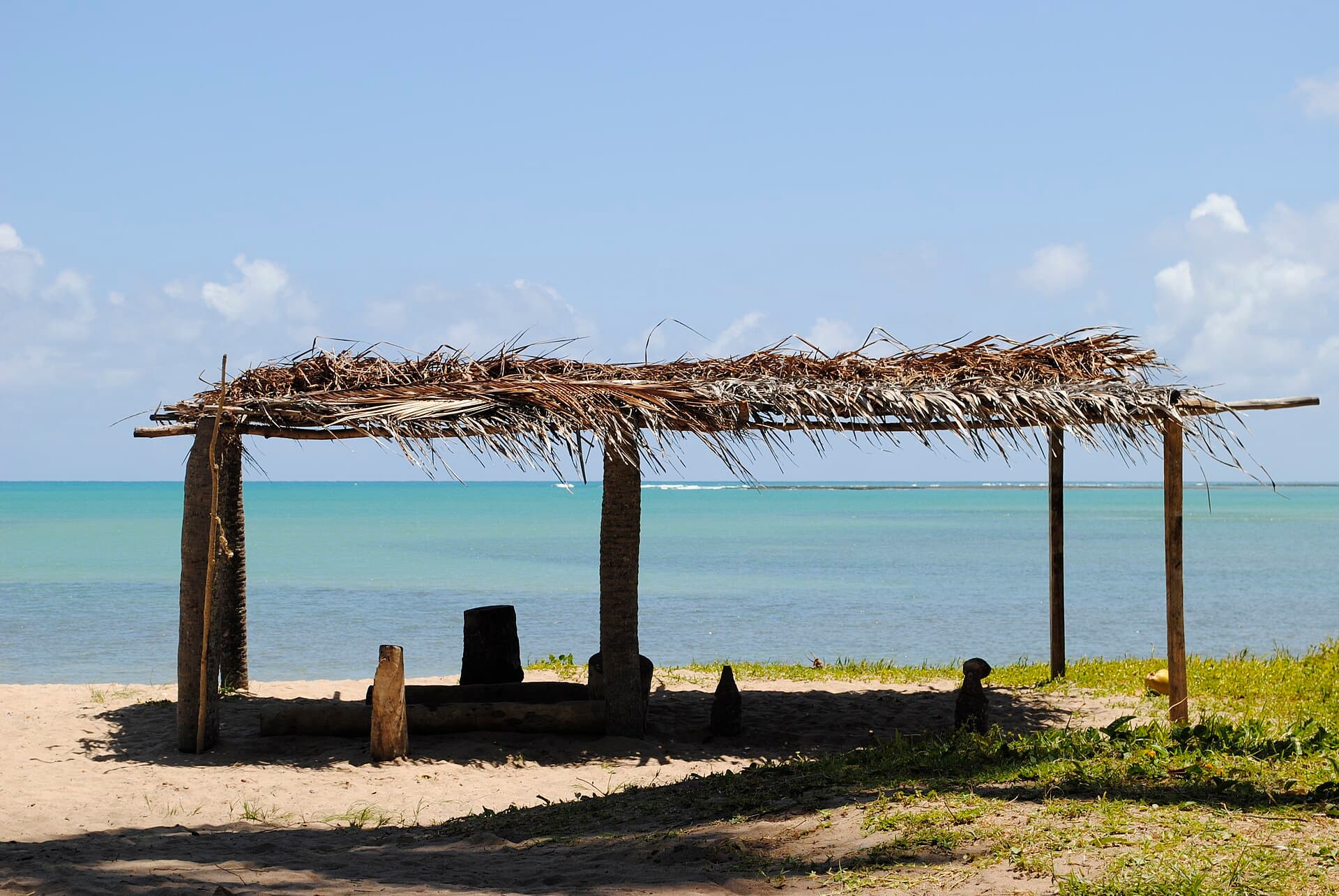 Caramanchão rústico de troncos e cobertura de palha seca sobre a areia de uma praia vazia, com o mar esverdeado atrás e a linha de recife quebrando ao longe