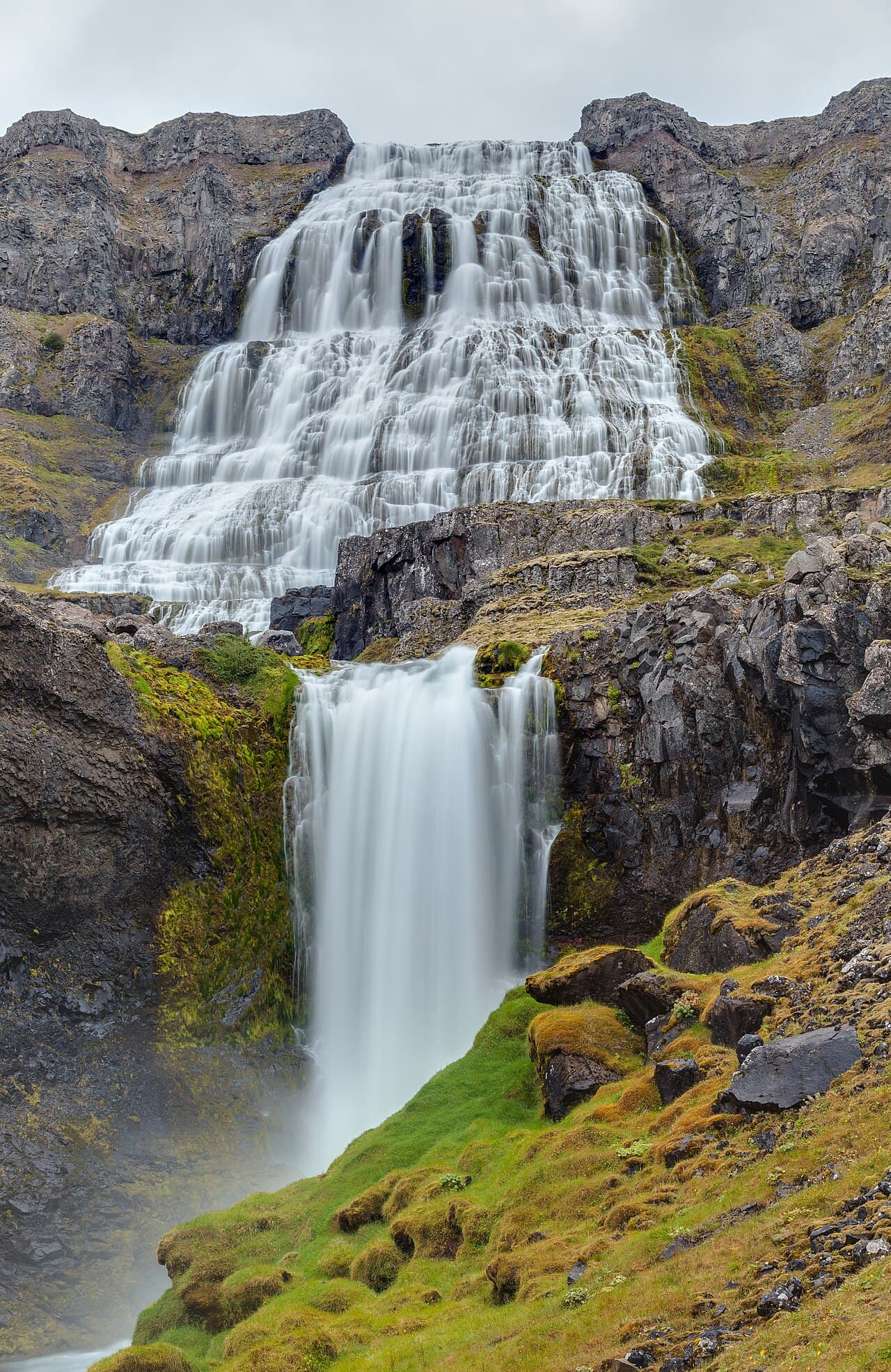 Cachoeira Dynjandi caindo em leque sobre degraus de rocha escura num fiorde dos Vestfirðir, na Islândia, com encosta verde ao redor